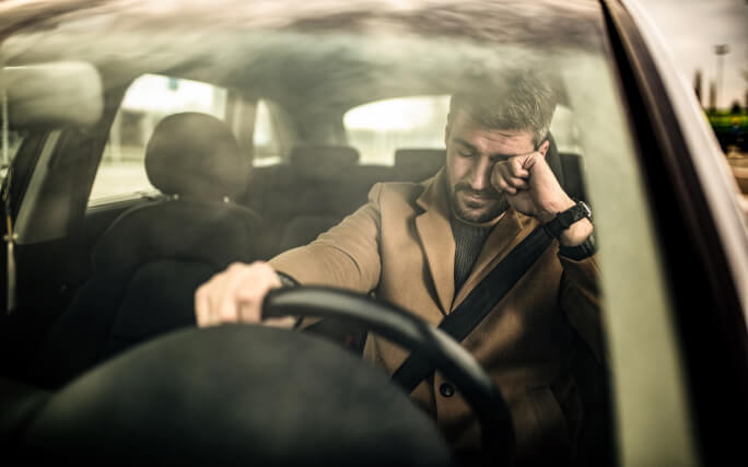 Homem dentro de um carro, em risco de adormecer ao volante.