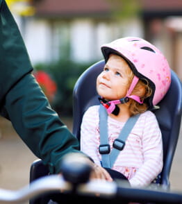 Uma menina sentada numa cadeira de bicicleta, com capacete e o cinto da cadeira apertado olha para um adulto. 