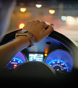 Mãos sobre o volante de um carro em marcha na estrada, sendo visíveis as luzes do painel do carro.  