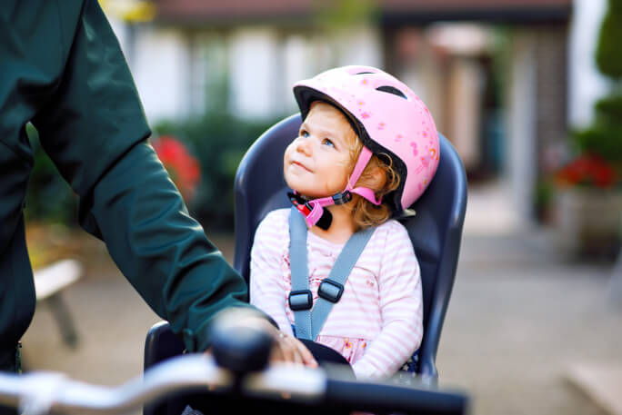 Uma menina sentada numa cadeira de bicicleta, com capacete e o cinto da cadeira apertado olha para um adulto. 
