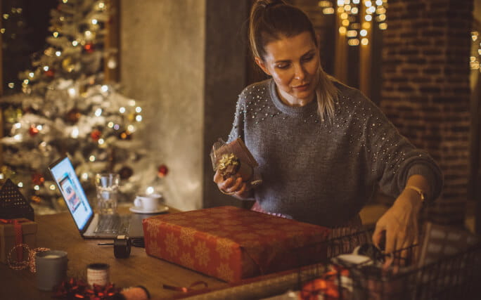 Uma rapariga numa sala com a árvore de Natal e presentes faz compras online num portátil.