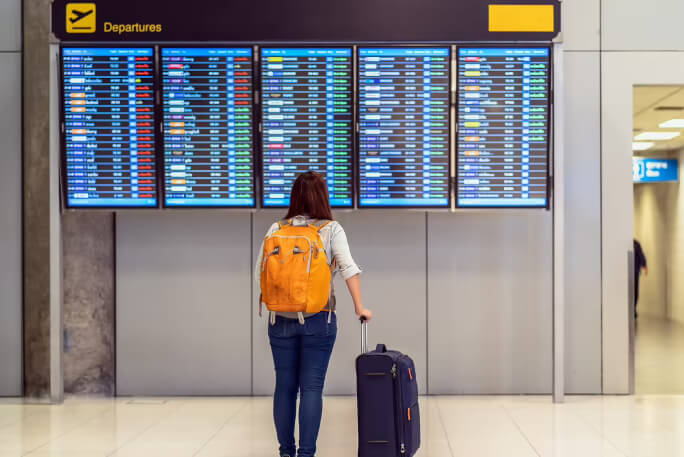 Uma rapariga de costas, num aeroporto, consulta o painel das partidas à procura da sua viagem.