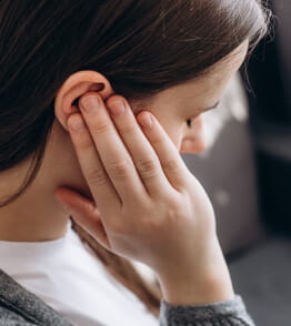 Mulher dentro de casa com ar de sofrimento aperta a orelha para tentar atenuar a dor de ouvido