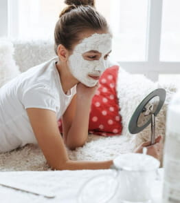Mulher jovem com máscara de cosmético sustentável aplicada no rosto, olha-se ao espelho  