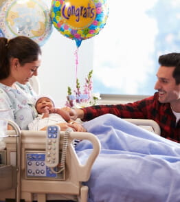 No quatro de um hospital, uma jovem mulher sentada na cama segura o primeiro filho ao colo e o pai faz-lhe uma festa. 