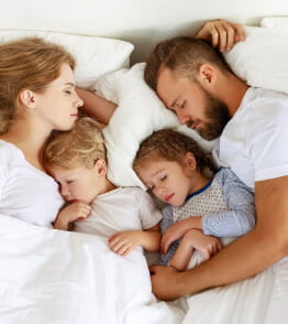Família de pai, mãe e dois filhos deitados a dormir na mesma cama.