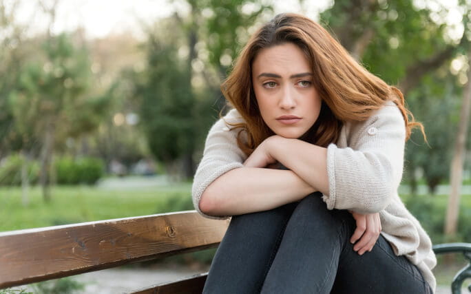 Jovem mulher sentada num banco de um jardim com um ar de grande preocupação. 